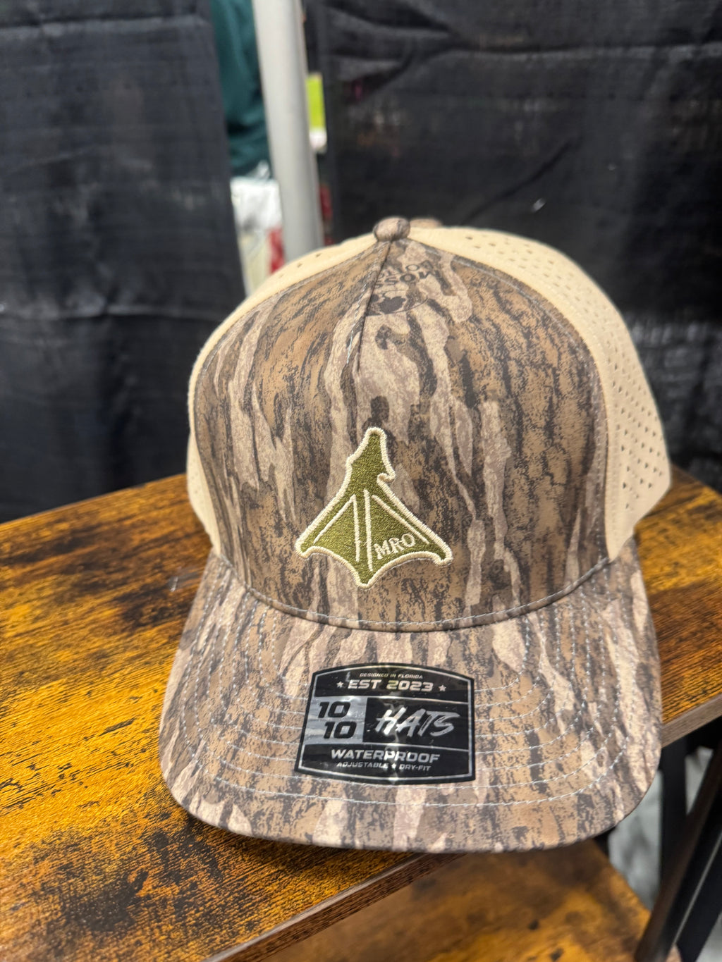 A camouflage trucker hat with a tan mesh back, featuring a green and tan webbed foot embroidered logo 