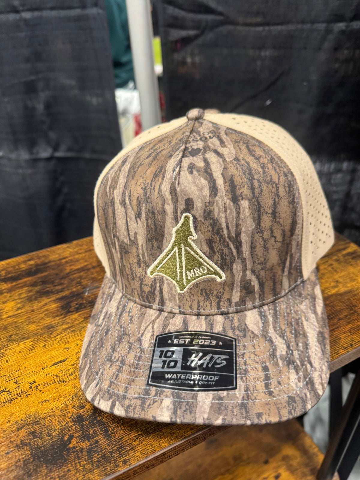 A camouflage trucker hat with a tan mesh back, featuring a green and tan webbed foot embroidered logo 